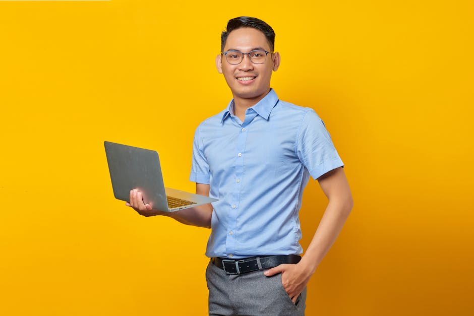 A confident young adult male with eyeglasses, holding a laptop against a vibrant yellow background, exuding a professional vibe.