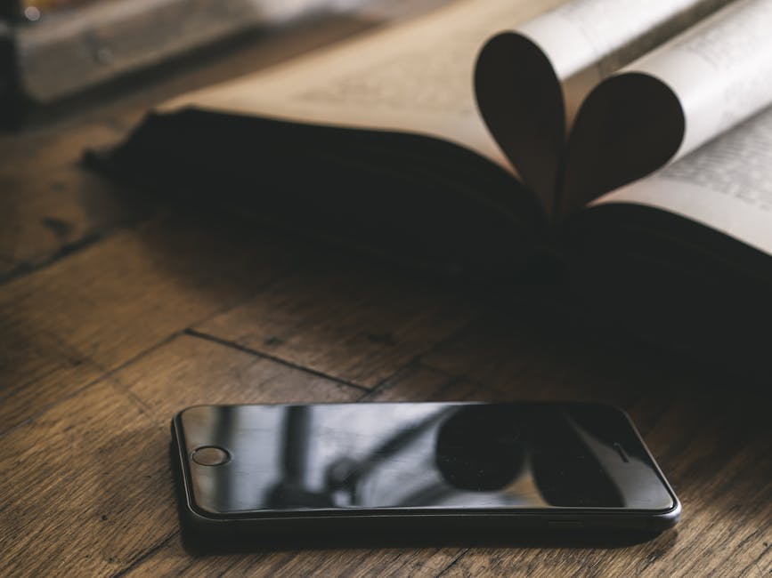 A smartphone rests on a wooden table beside an open book with pages forming a heart.