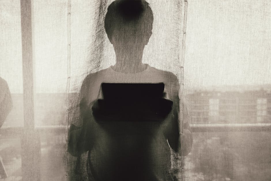 A silhouette of a person using a tablet device behind a curtain indoors, creating a mysterious ambiance.