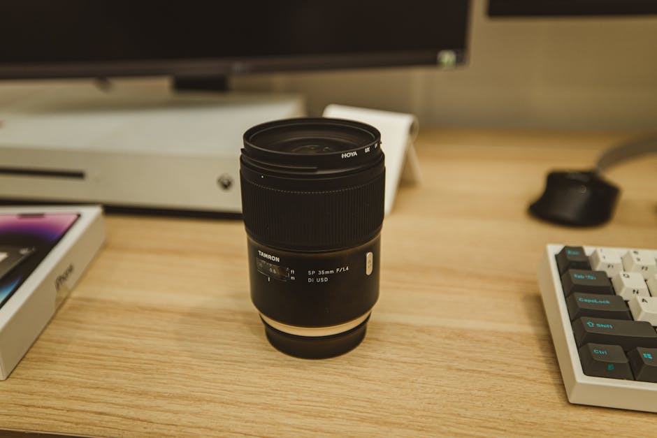 A Tamron SP 35mm camera lens placed on a wooden desk with office equipment and a monitor.