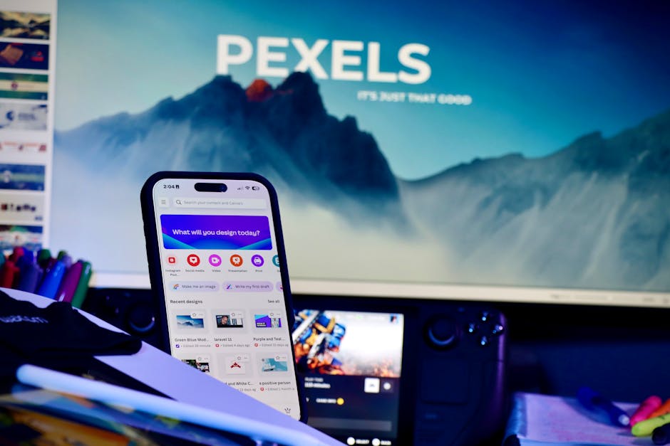 A creative workspace with a smartphone displaying design apps and a large monitor in the background.