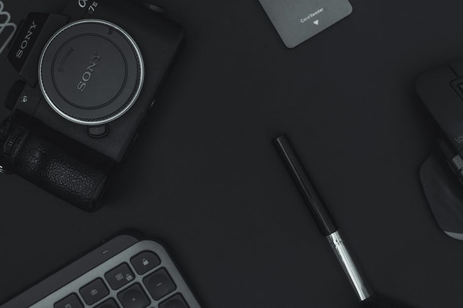 Top view of a minimalist desk setup featuring a camera, keyboard, pen, and card reader on a dark surface.
