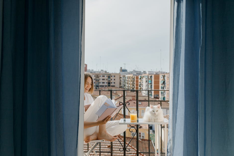 A woman enjoys reading on her balcony with a white cat and a glass of juice.