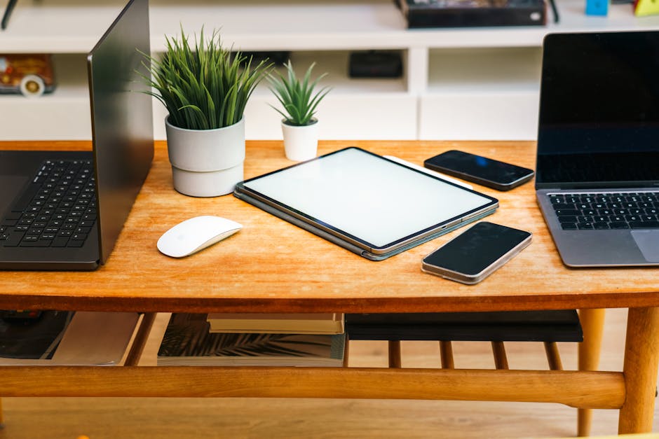 A stylish home office desk with laptops, tablet, smartphones, and potted plants.