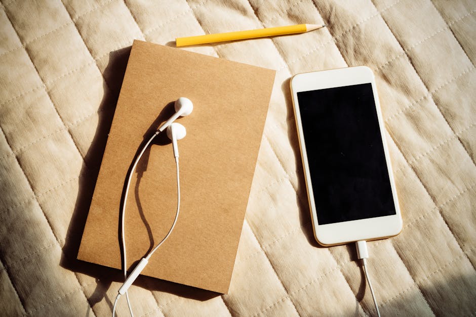 A top-view shot of a smartphone, earbuds, notebook, and pencil on a bedspread.